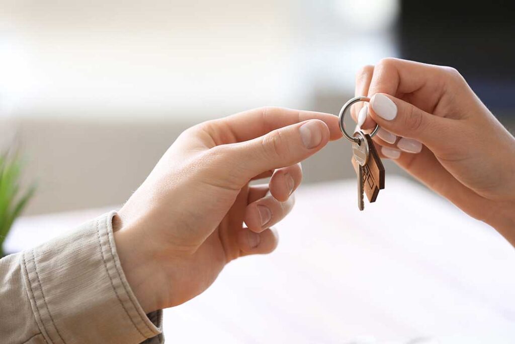 A person’s hand passing house keys over a contract on a table with real estate documents and a pen nearby.