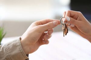 A person’s hand passing house keys over a contract on a table with real estate documents and a pen nearby.